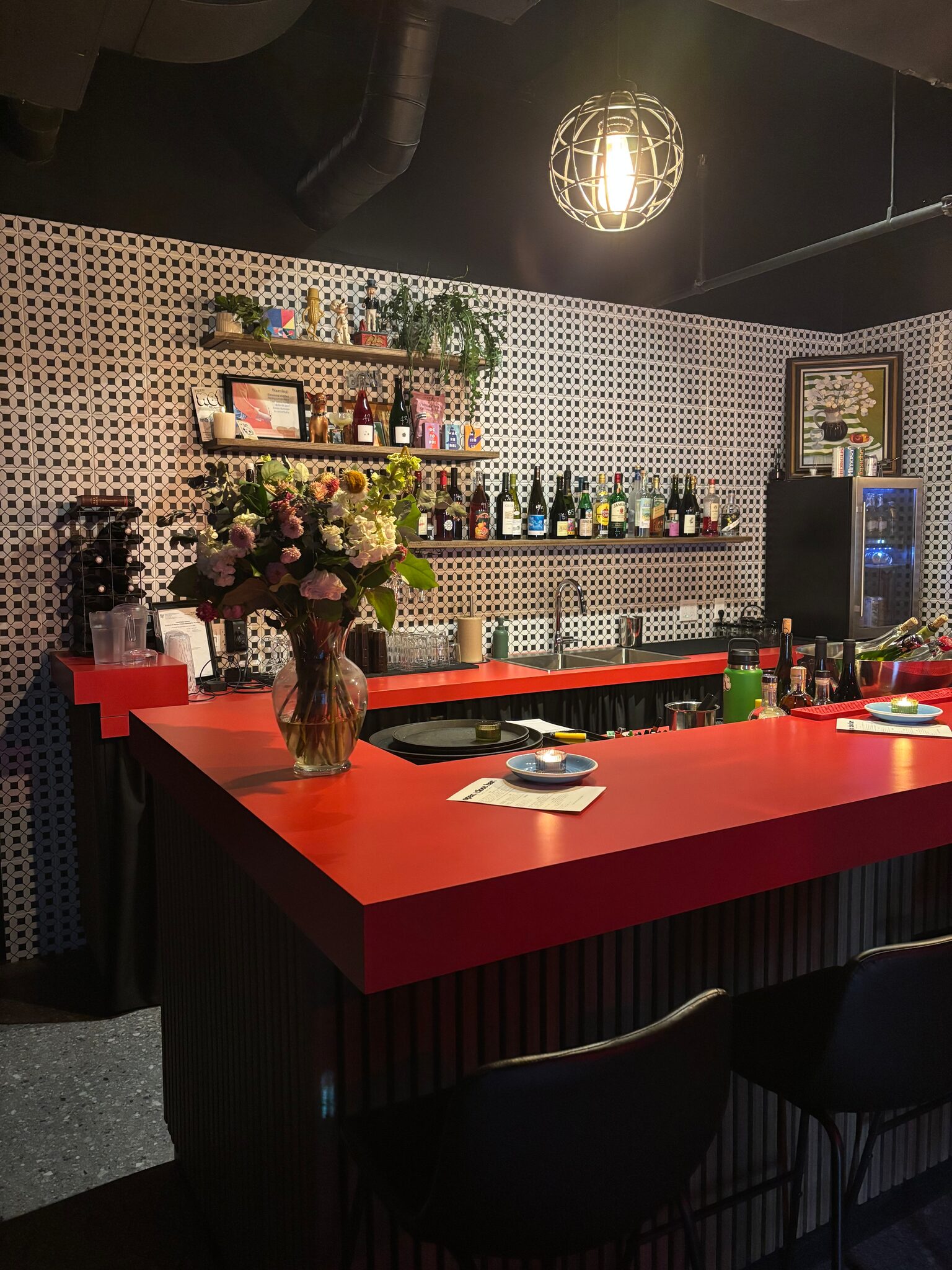 the red bar top with flowers and candles, black stools, patterned tile and shelves of wine under a hanging lamp